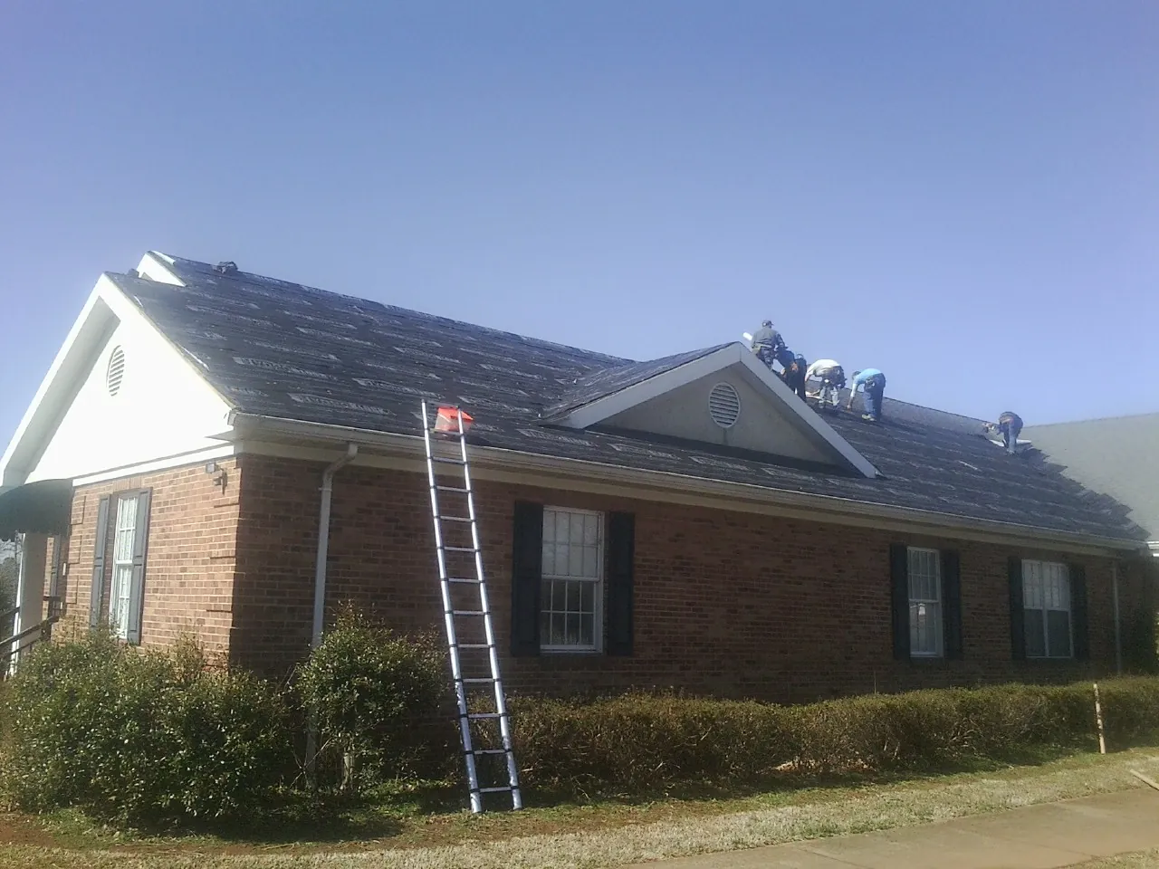 Crew tearing shingles off a brick ranch roof with a ladder set — Metal Roof Repair work by our crew in Hye